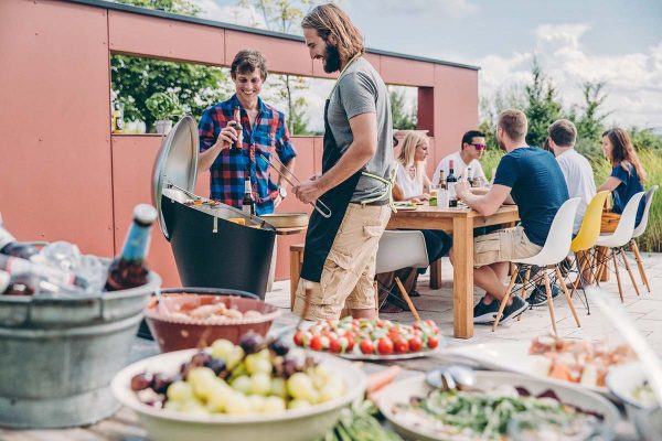 Två män står och grillar på Höfats Cone Kolgrill under grillfest med sällskap i bakgrunden